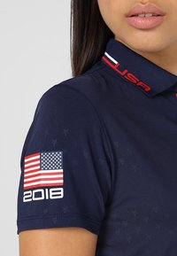 Marinblå pikétröja med röda detaljer, texten USA på kragen, och ett märke med den amerikanska flaggan och "2018" på vänster ärm.