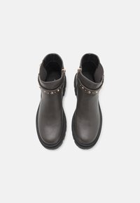 Botas de ankle de cuero negro con suela plana, adornadas con tachuelas doradas en la correa y un diseño bajo. Textura suave con detalle de cremallera.