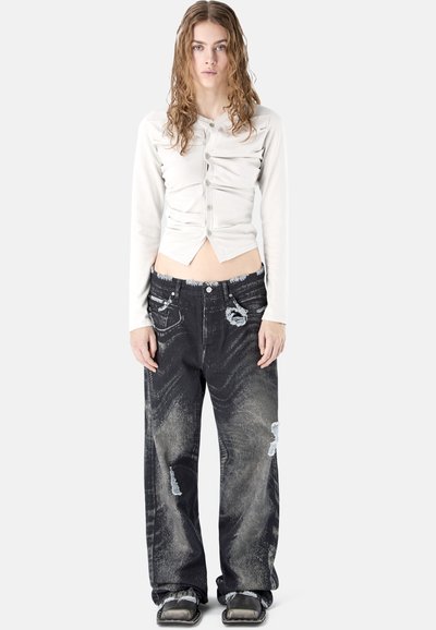 Mujer joven con cabello ondulado que lleva una blusa blanca de manga larga con botones y fruncidos, y pantalones vaqueros negros holgados con desgaste y zapatos de punta cuadrada.