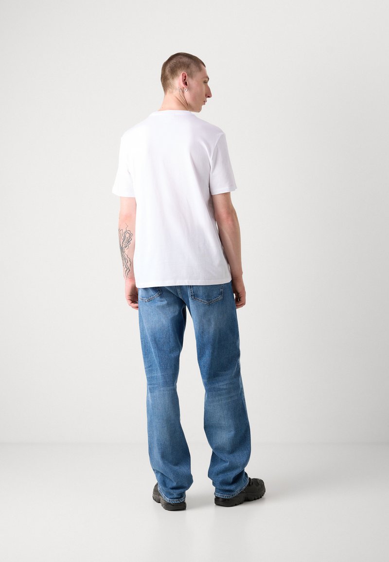 Biała koszulka z krótkim rękawem w połączeniu z luźnymi niebieskimi dżinsami, z kieszeniami i swobodnym krojem. U dołu widoczne czarne obuwie.