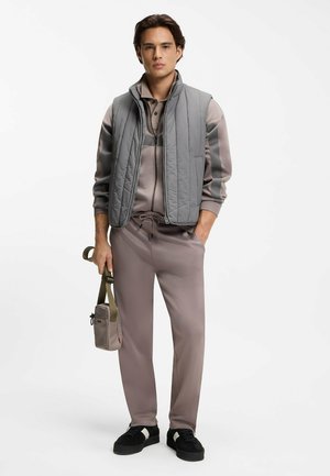 Young man wearing a mauve tracksuit and gray quilted vest, holding a small beige crossbody bag with olive strap, standing with one hand in pocket.