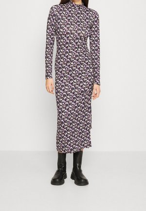 Femme portant une robe midi cintrée à manches longues avec un petit motif floral violet, rose et blanc, associée à des bottines noires.