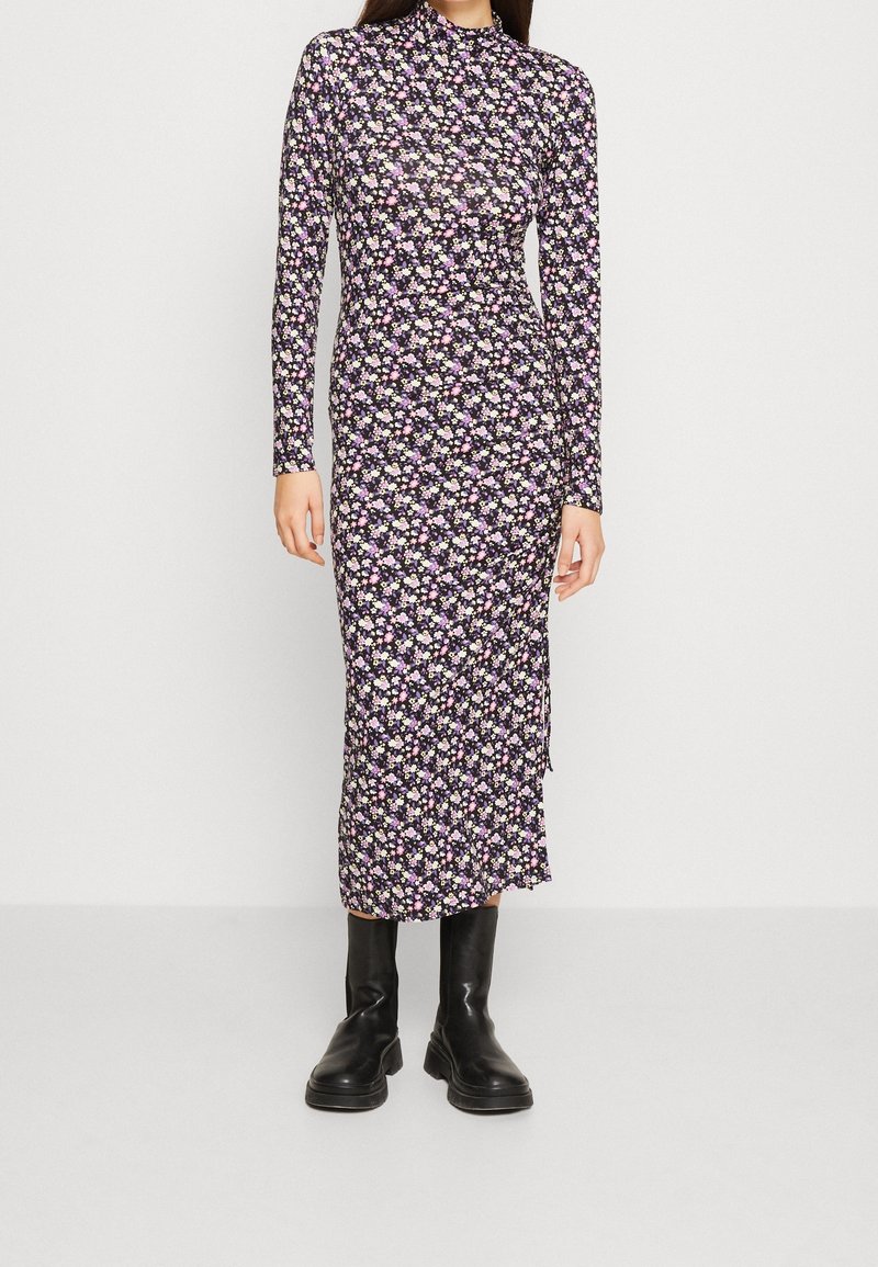 Femme portant une robe midi cintrée à manches longues avec un petit motif floral violet, rose et blanc, associée à des bottines noires.