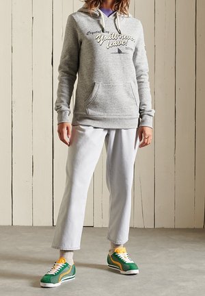 Person trägt eine graue Kapuzenjacke mit Text, lichtfarbene, gekürzte Hosen, beige Socken und grüngelbe Turnschuhe und steht auf einem Betonboden vor hölzernen Wandpaneelen.