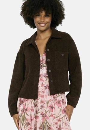 Braune, flauschige Jacke mit Kragen, vorderen Knöpfen und einer Brusttasche, getragen über einem blumengemusterten rosa Kleid.