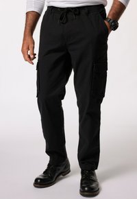 Zwarte cargobroek met elastische tailleband en trekkoord. Bevat zijzakken en heeft een relaxte pasvorm. Draagt met zwarte leren schoenen.