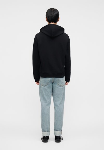 Μαύρο φούτερ με κουκούλα και άνετη εφαρμογή, συνδυασμένο με ανοιχτό μπλε τζιν. Το τζιν έχει γυρισμένα μανίκια και κλασικό σχέδιο με πέντε τσέπες.