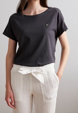 Donkergrijze katoenen T-shirt met korte opgerolde mouwen en een ronde halslijn; heeft een kleine geborduurde palmboom op de borst. Gecombineerd met witte gestructureerde broeken.