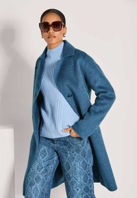 Vrouw met grote donkere zonnebril, blauwe turtlenecktrui, blauwe geblokte jeans en een blauwe wollen jas met één hand in haar zak.