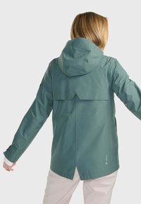 Veste verte imperméable avec capuche, ourlet arrière incurvé et poignets en velcro. Fabriquée en matériau GORE-TEX avec une texture lisse et mate.