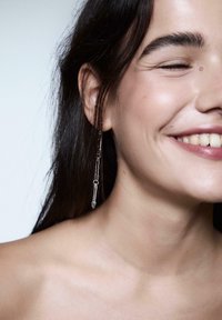 Giovane donna dai capelli scuri che sorride, indossando un grande orecchino a catena argentata, mostrando i denti e con gli occhi chiusi e lievi rughe alle guance.