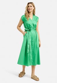 Grünes, knielanges, kurzärmliges Kleid mit V-Ausschnitt, taillierten Bund und seitlichen Taschen; aus glattem Stoff in einfarbiger Ausführung.