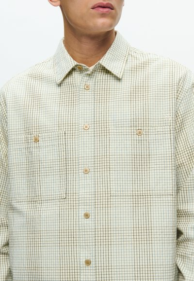 Chemise boutonnée en beige clair avec un motif à carreaux en brun foncé et bleu. Elle présente deux poches poitrine et des boutons en bois.