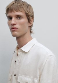 Light beige linen shirt with a collar, front placket, and two chest pockets. Features brown buttons and a subtle, textured weave.