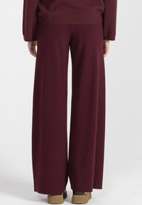 Osoba w szerokich burgundowych spodniach dzianinowych i dopasowanej bluzie, pokazana od tyłu, stojąca na białym tle.