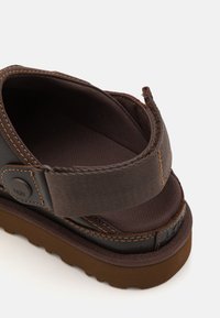 Bruine sandaal met een bovenwerk van textuurstof, gevoerde banden en een bruine rubberen zool. Decoratieve stiksels langs de randen. UGG logo detail.