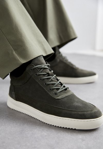 Dark gray suede sneakers with white soles worn with olive green pants, shown on a light gray floor.