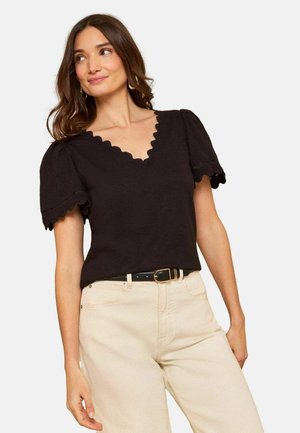 Moteris su banguotais rudais plaukais dėvi juodą viršų su raukšlėtais V formos iškirpimu ir pūstomis rankovėmis bei smėlio spalvos kelnes su juodu diržu, žvelgia šiek tiek į kairę.