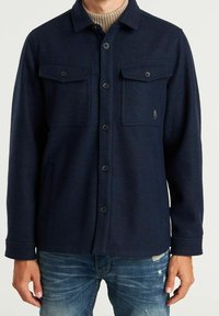 Mann trägt eine marineblaue Jacke mit Knöpfen über einem beigen, gerippten Rollkragenpullover und abgenutzten blauen Jeans, gezeigt von den Schultern bis zur Hüfte.