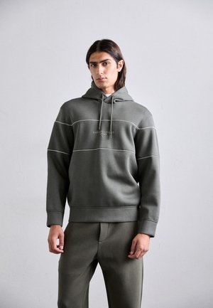 Hanorac cu glugă de culoare verde măsliniu, cu accente de cusături albe și buzunar tip kangaroo; asortat cu pantaloni care sunt din același material moale.