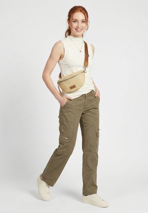 Smiling woman with red hair wears sleeveless white top, olive cargo pants, white sneakers, and crossbody beige bag, standing with hands in pockets.