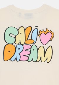 Kremna majica s poudarjenim, barvitim grafičnim besedilom: "CALI", "HEART" in "DREAM" v različnih pisavah in oblikah, podobnih mehurčkom.