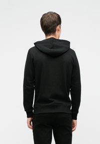 Homme portant un sweat à capuche noir et un pantalon noir, debout de dos devant un fond uni gris clair.
