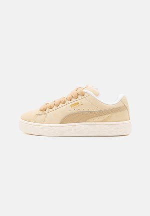 Beige mocka sneakers med texturerad yta, rund tå, platta beige snören, vita detaljer på hälen och ett mönstrat gummisul.