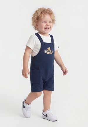 Malé dieťa kučeravými vlasmi sa usmieva, kráča, má na sebe biele tričko, tmavomodré nohavice s trakmi Moschino s logom medveďa a biele tenisky Nike.