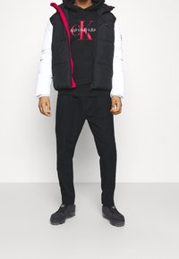 Gilet piumino nero con maniche bianche e dettagli rossi sopra un felpa nera, abbinato a pantaloni neri e calzature scure. Tessuto liscio, vestibilità casual.