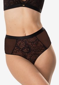Kobieta w czarnym koronkowym bieliźnie i pasującym biustonoszu, prezentująca środkową część ciała i górne uda na tle białym.