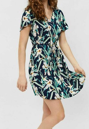 Eine Frau trägt ein kurzärmliges, marineblaues Blumenkleid mit weißen und grünen Blättern und hält den Saum des Kleides leicht hoch.