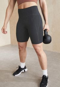 Černé sportovní kraťasy s vysokým pasem a švy, doplněné černými sportovními botami a bílými kotníkovými ponožkami, které drží kettlebell.