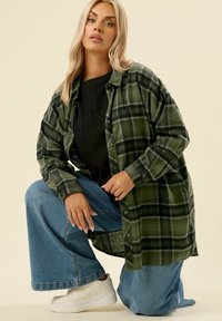 Camicia a flanella a quadri verdi, con bottoni, indossata sopra una maglietta nera, abbinata a jeans blu a gamba larga e scarpe da ginnastica bianche.