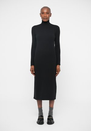 Hosszú ujjú, fekete kötött ruha magas nyakkivágással, karcsú szabással és egyenes sziluettel, szürke zoknikkal és fekete lomha cipőkkel, arany részletekkel párosítva.