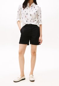 Hvid skjorte med knapper og sejlbådsmønster, parret med sorte bomuldsbukser og cremegule loafers med guldfarvede detaljer.