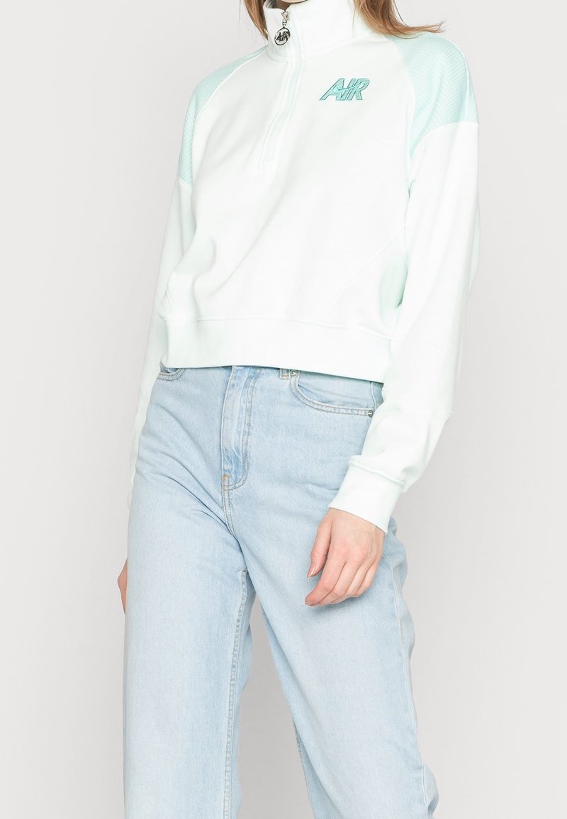 Personne portant un jean taille haute bleu clair et un sweat zippé blanc avec des accents bleu clair et le logo "AIR" sur la poitrine.
