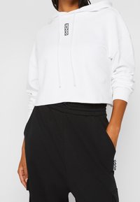 Woman wearing a white cropped hoodie with "HUGO" logo and black drawstring pants with hand in pocket, shown from chest to hips.