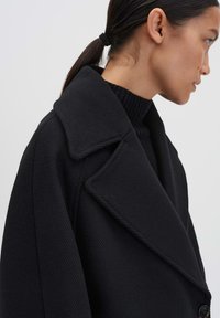 Cappotto in misto lana nero con ampio rever, tessuto texture e dettagli con bottoni sui lati. Il modello indossa un dolcevita nero a coste sotto.