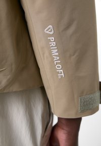 Beige jacket sleeve featuring Primaloft® logo embroidered in white. Jacket material appears smooth with a subtle sheen, and includes a gray Velcro accent.