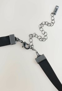 Schwarzer Nylonriemen mit metallischen Beschlägen, der mit strukturierten Enden und einer Kettenverlängerung versehen ist. Enthält einen Karabinerhaken für sicheren Halt.