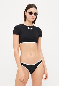 Fekete úszófelső rövid ujjakkal és fehér díszítéssel, gyémánt logóval. Hozzá illő bikini alsóval fehér akcentussal.