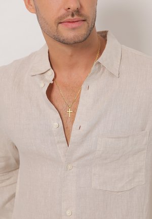 Chemise en lin beige clair avec un col boutonné, une poche poitrine et des coutures visibles. Portée avec un collier en or en forme de croix.