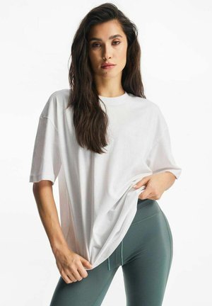 Kobieta o długich ciemnych włosach, ubrana w luźną białą koszulkę i zielone legginsy z wysokim stanem, poprawiająca koszulkę w talii.