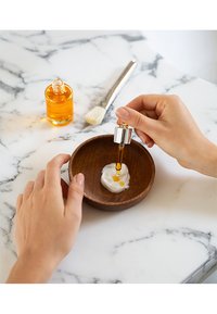 Träskål med vit grädde och apelsinolja, hand som applicerar oljan med pipett. Marmor yta, pensel i närheten.