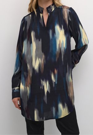 Tunique à manches longues avec col montant et motif abstrait de coups de pinceau bleus, noirs, beiges et bruns, portée avec un pantalon noir.