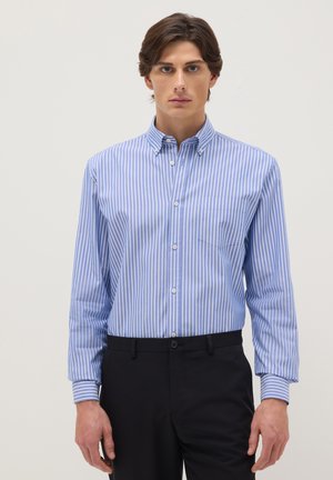 Homme portant une chemise à rayures verticales bleu et blanc boutonnée et rentrée dans un pantalon noir, debout devant un fond uni.