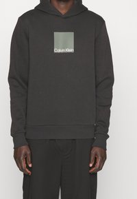 Man draagt een zwarte Calvin Klein hoodie met een groen vierkant logo op de borst en bijpassende zwarte broek, staand tegen een witte achtergrond.