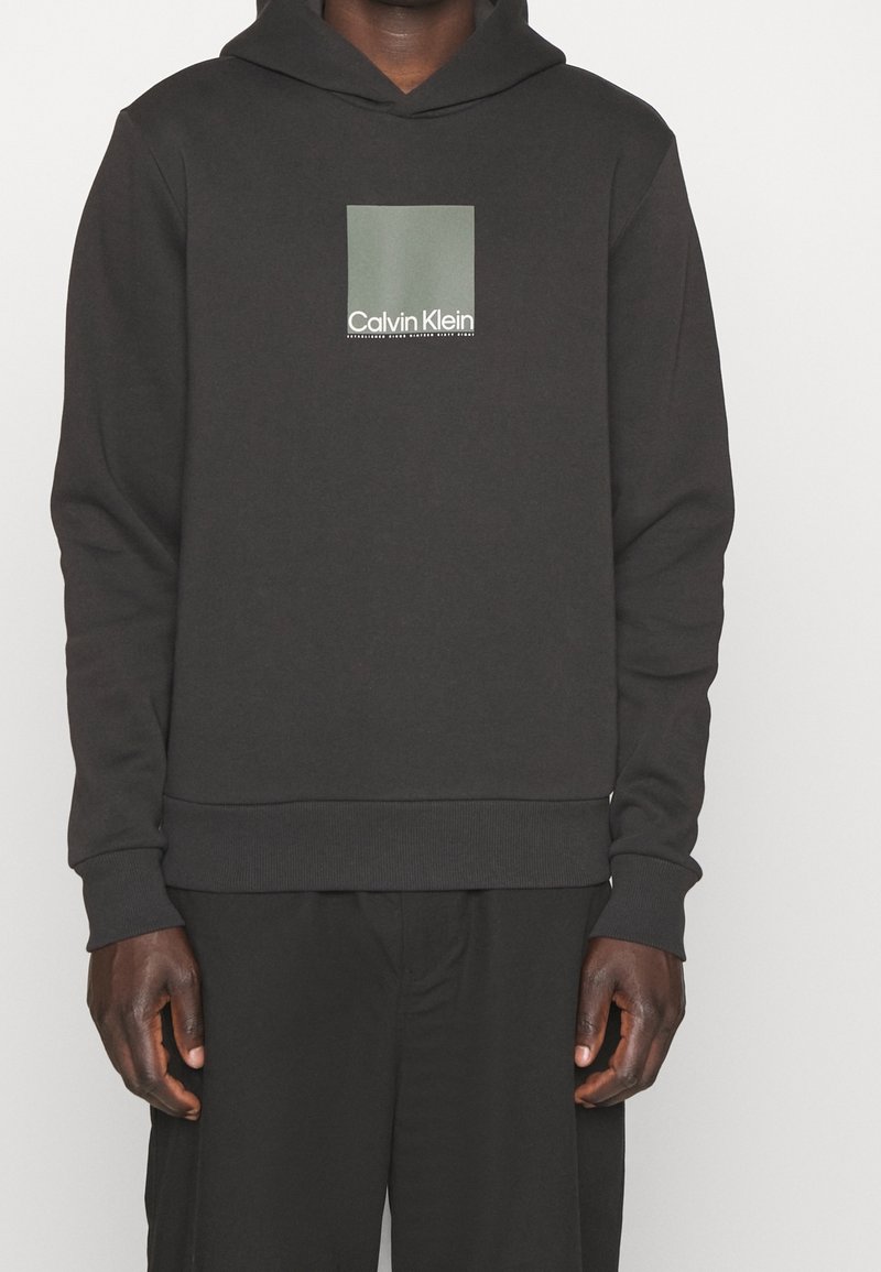 Man draagt een zwarte Calvin Klein hoodie met een groen vierkant logo op de borst en bijpassende zwarte broek, staand tegen een witte achtergrond.