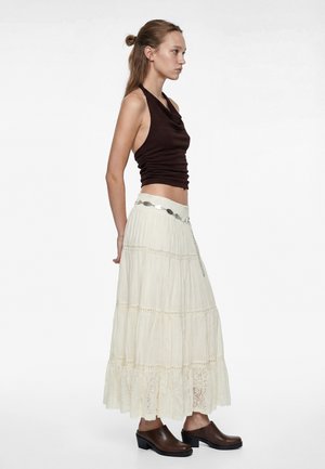 Joven mujer con una blusa sin mangas de color marrón oscuro, falda larga de encaje con volantes color blanco roto, cinturón plateado y zuecos marrones de tacón, de pie de perfil.